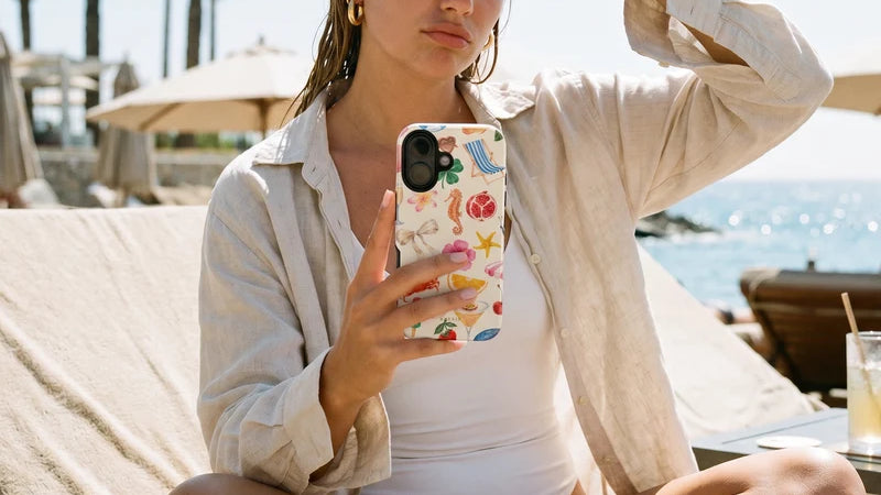 A woman sitting on a beach lounge chair outdoors, holding a phone with a colorful tropical seashell and coral print case. She is wearing a white one-piece swimsuit and a light beige button-up shirt.
