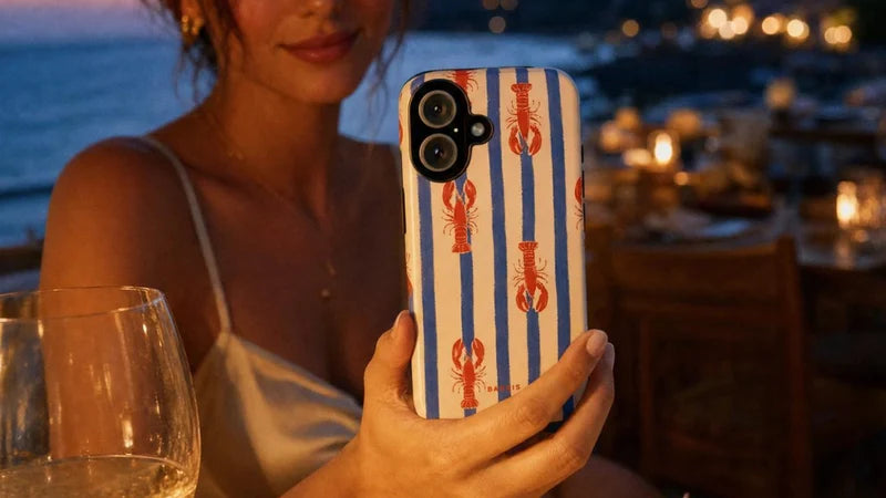 A woman at a dinner table holds up a smartphone with a blue and white striped case decorated with red lobster illustrations. A cooked lobster, a glass of white wine, and a plate with a lemon slice are on the table in front of her, with a scenic coastal town at dusk in the background.