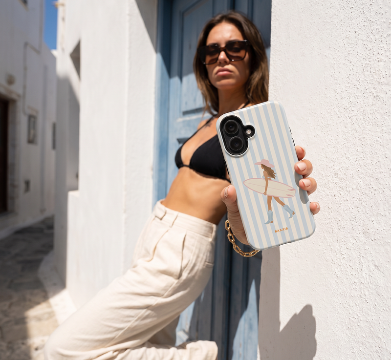 A woman in a black bikini top and beige pants holds up a light blue and white striped phone case featuring a cartoon illustration of a woman with a surfboard. The background shows a sunny, sunlit white wall and a blue door.