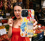 A woman in a red dress holds up a phone case featuring a colorful 'Ciao Amore' Sicilian collage design with illustrations of chili peppers, waves, and local landmarks, set against a busy market background filled with red chilies.