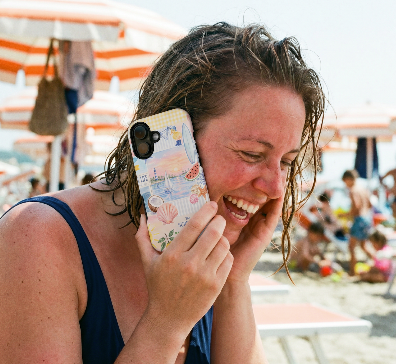 A woman with wet hair smiling while holding a phone to her ear on a beach. The phone has a pastel multicolor illustration case with a camera cutout.