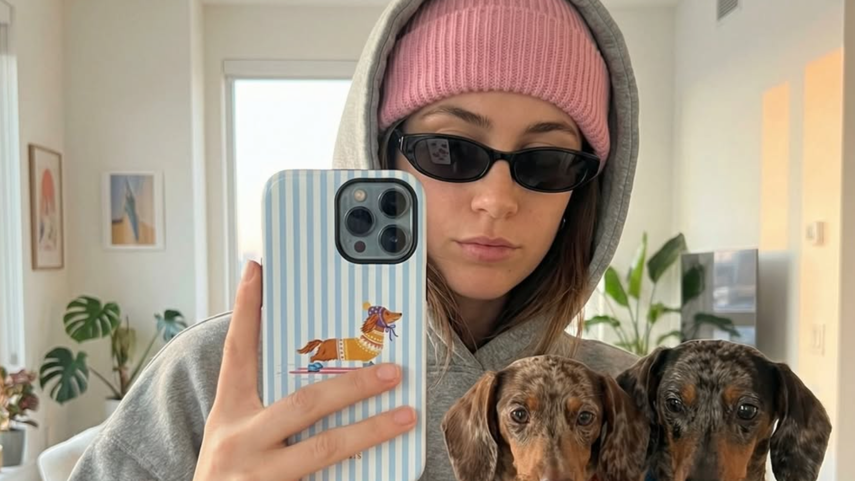 A person taking a mirror selfie indoors, wearing a gray hoodie, pink beanie, and black sunglasses, holding a light blue and white striped phone case with a cartoon dachshund graphic. Two dachshunds wearing colorful sweaters are held in the person's arms.