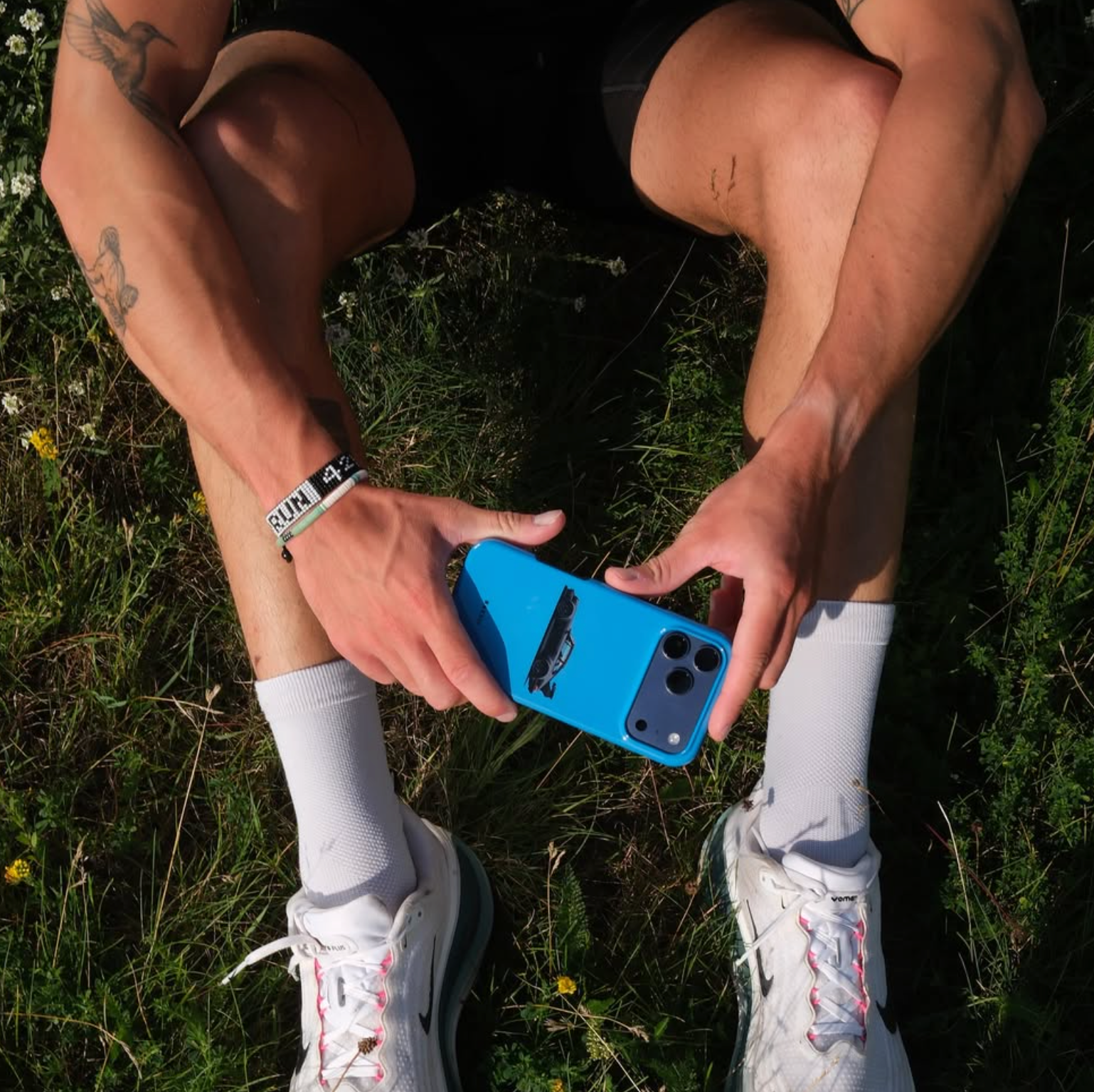 A top-down view of a person outdoors in grass, holding a blue smartphone case with a black strap. The person is wearing white socks, white running shoes with pink laces, and a black-and-white wristband that reads 'RUN 24'.