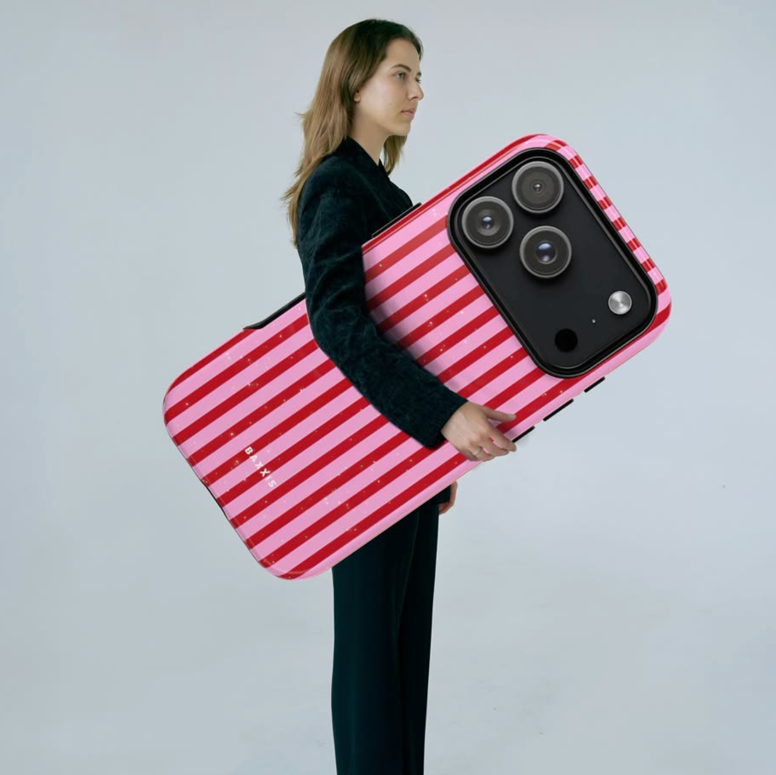A woman in a black outfit holds a very large smartphone case with bold pink and red diagonal stripes. The case is designed to look like a modern smartphone, featuring a black camera lens surround and a small white logo near the bottom left.
