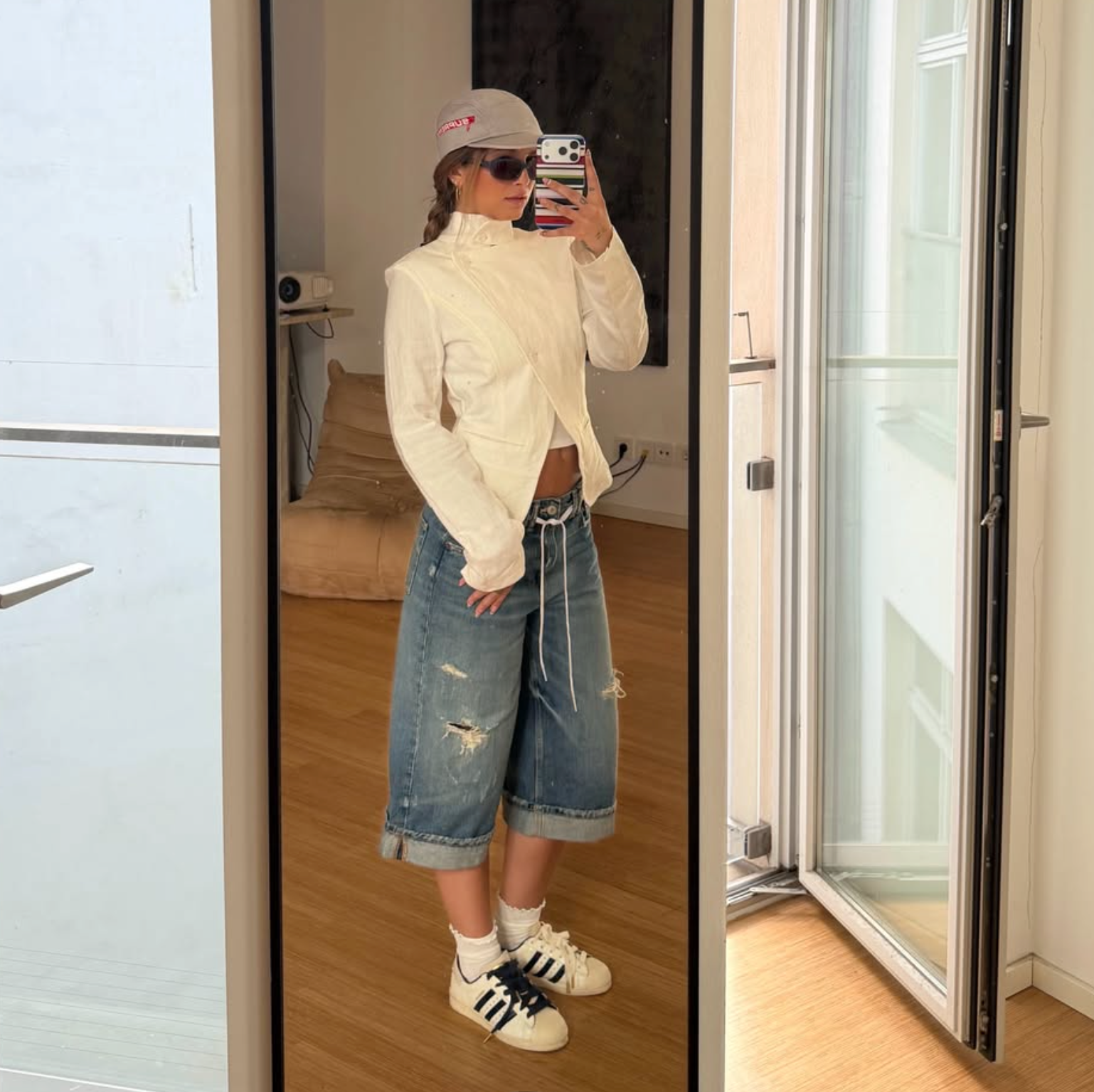 A person taking a mirror selfie in an indoor room, wearing a cream cropped jacket, distressed blue denim shorts, white socks, and white sneakers with black stripes. The background shows a wooden floor, a sofa, and glass doors.