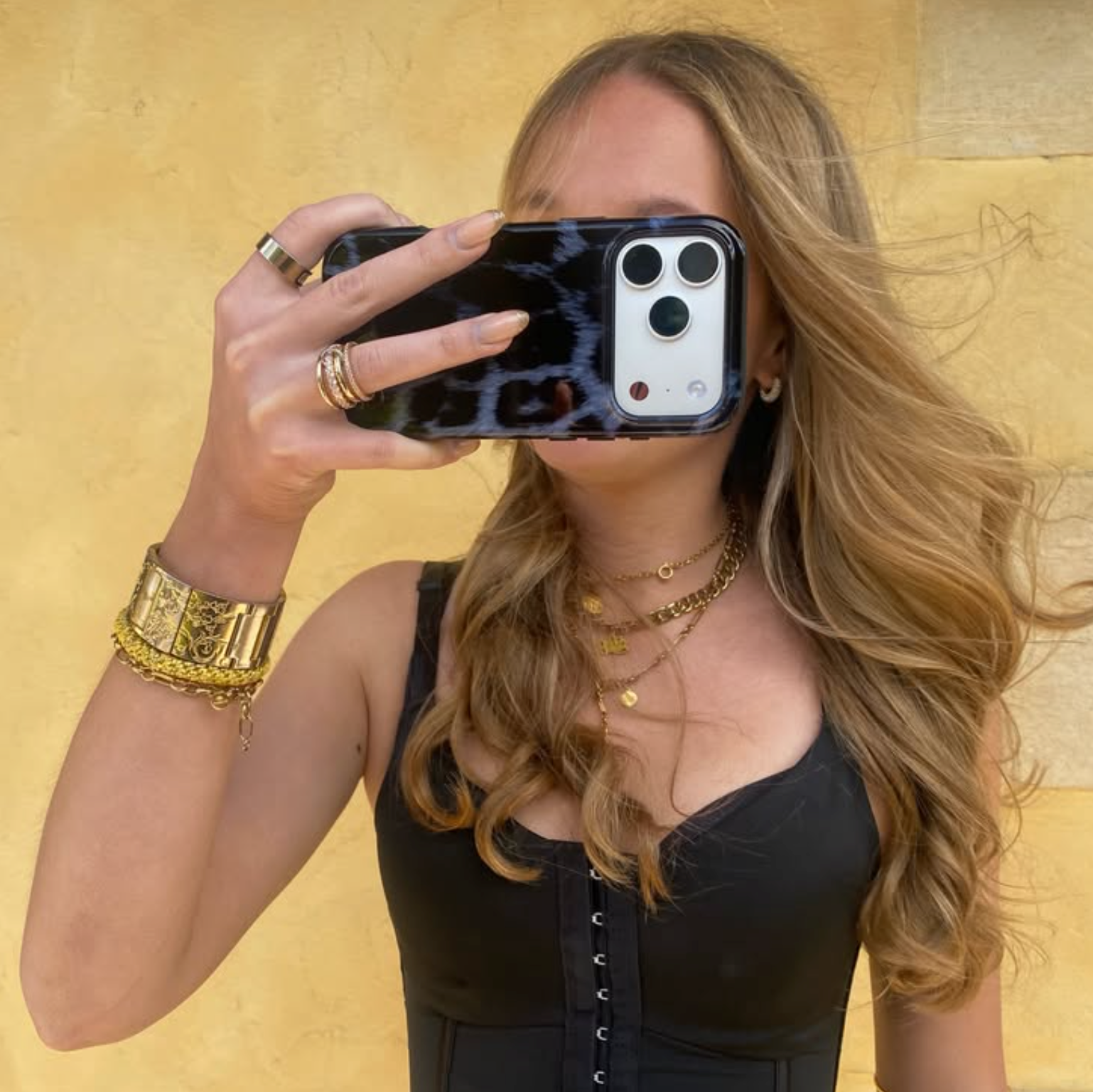 A woman taking a mirror selfie wearing a black lace-up top, gold jewelry, and a black leopard-print phone case against a warm yellow stone wall.