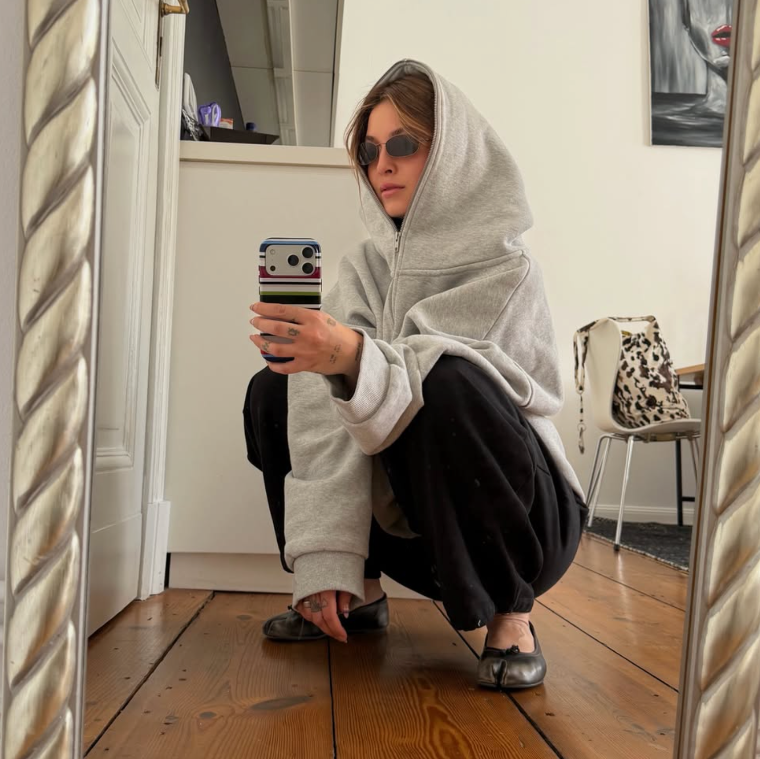 A person takes a mirror selfie in a home setting, wearing an oversized light gray hoodie, black pants, and black flat shoes. The background shows a wooden floor, white walls, and a chair with a leopard print bag.